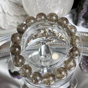 May include: A bracelet made of clear quartz beads with gold inclusions. The beads are round and have a smooth, polished finish. The bracelet is displayed on a clear stand.