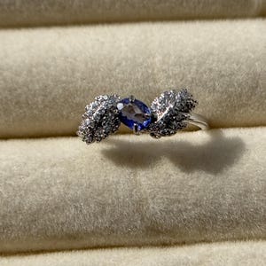 May include: A silver ring featuring a central oval-cut, deep blue gemstone, flanked by two stylized leaf designs encrusted with small, sparkling white stones. The ring is set against a soft, light-colored background.