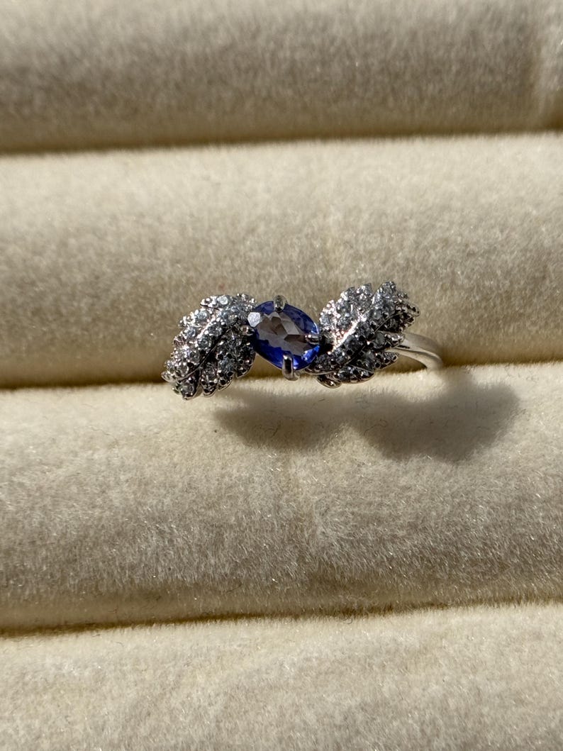 May include: A silver ring featuring a central oval-cut, deep blue gemstone, flanked by two stylized leaf designs encrusted with small, sparkling white stones. The ring is set against a soft, light-colored background.