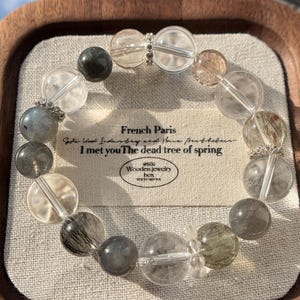 May include: A beaded bracelet featuring a variety of clear, gray, and amber-colored stones. The bracelet includes a tag with the text "French Paris" and "I met you The dead tree of spring". The bracelet is displayed on a linen surface.