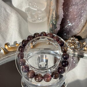 May include: A beaded bracelet with dark purple and brown stones. The bracelet is displayed on a clear, tiered stand, reflecting on a mirrored surface. The beads appear to be approximately 1 cm in diameter.