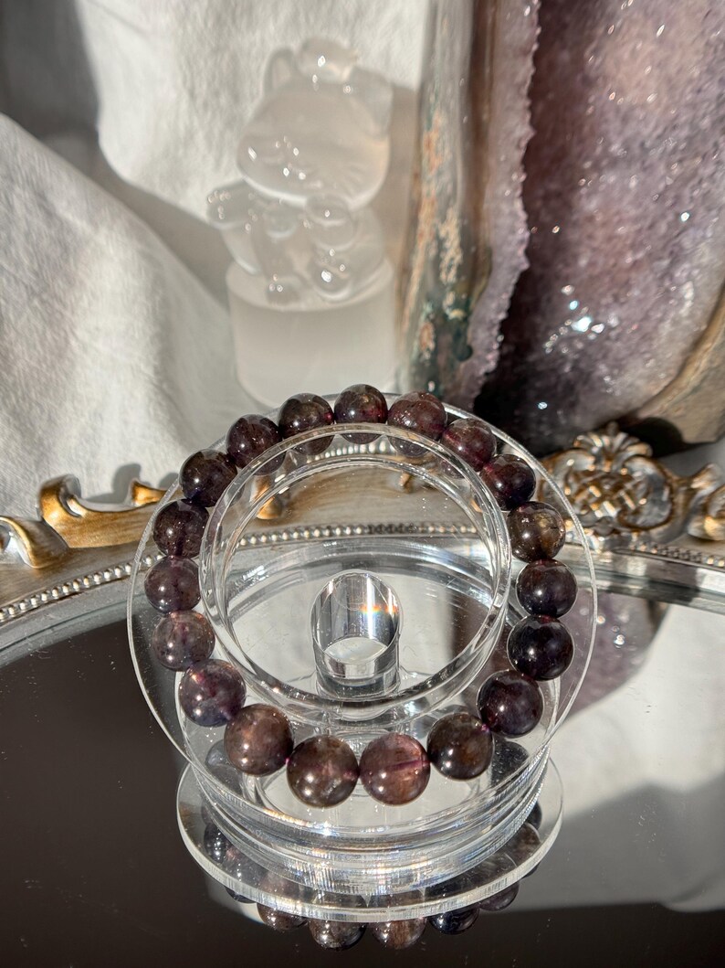 May include: A beaded bracelet with dark purple and brown stones. The bracelet is displayed on a clear, tiered stand, reflecting on a mirrored surface. The beads appear to be approximately 1 cm in diameter.