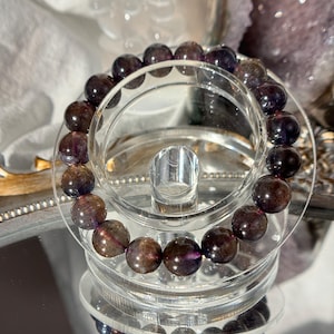 May include: A bracelet of polished, round, dark purple and brown beads. The bracelet is presented on a clear, circular stand, reflecting on a mirrored surface. The beads have a subtle, natural sheen.