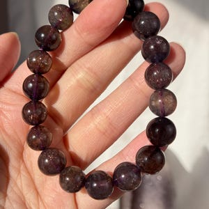 May include: A bracelet made of large, round, dark purple beads with a shimmering, translucent appearance. The beads are strung together, forming a piece of jewellery. The bracelet is held in a hand.