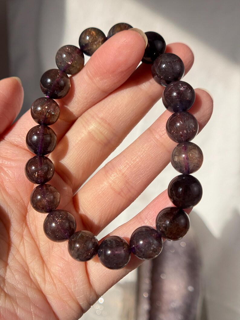 May include: A bracelet made of large, round, dark purple beads with a shimmering, translucent appearance. The beads are strung together, forming a piece of jewellery. The bracelet is held in a hand.