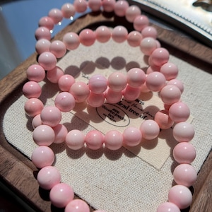 May include: Three pink beaded bracelets. The bracelets are made of round, light pink beads. The beads are strung together on an elastic band. The bracelets are displayed on a wooden tray with a small tag that reads "Wooden jewelry box".