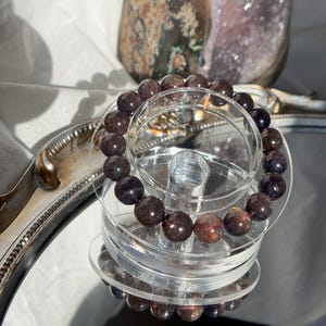 May include: A bracelet made of dark burgundy and brown gemstone beads, displayed on a clear, tiered stand. The bracelet is reflected on a mirrored surface. A decorative glass object is visible in the background.