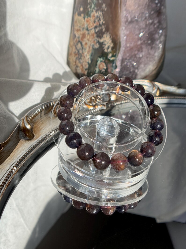 May include: A bracelet made of dark burgundy and brown gemstone beads, displayed on a clear, tiered stand. The bracelet is reflected on a mirrored surface. A decorative glass object is visible in the background.