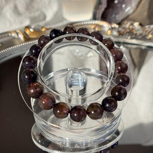 May include: A bracelet of dark red and black gemstone beads. The bracelet is displayed on a clear acrylic stand, reflecting on a mirrored surface. The beads are polished and spherical in shape.