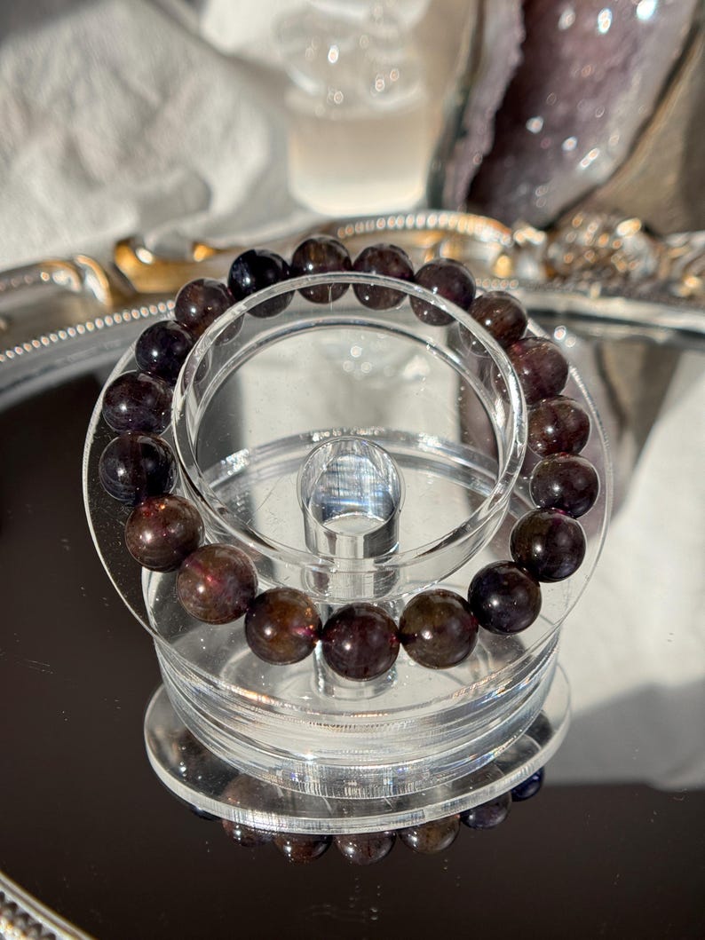 May include: A bracelet of dark red and black gemstone beads. The bracelet is displayed on a clear acrylic stand, reflecting on a mirrored surface. The beads are polished and spherical in shape.