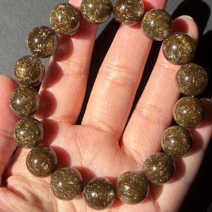 May include: A bracelet composed of spherical, translucent, golden-brown beads. Each bead displays a shimmering, fibrous interior, giving a unique, textured look. The bracelet is held in a hand, highlighting the natural beauty of the gemstone.