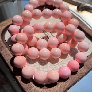 Può includere: Una collezione di braccialetti di perline rosa. Le perline sono rotonde e hanno un aspetto liscio e lucido. I braccialetti sono esposti su un vassoio di legno con un rivestimento in tessuto chiaro. Il colore delle perline varia leggermente.