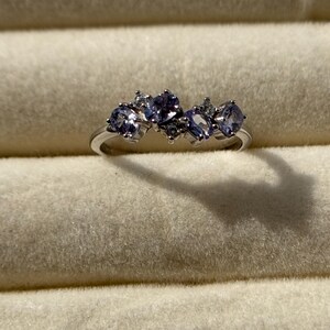 May include: A silver ring featuring five oval-shaped, light purple gemstones. The ring is set with smaller, clear accent stones. The ring is displayed on a light beige, textured surface.