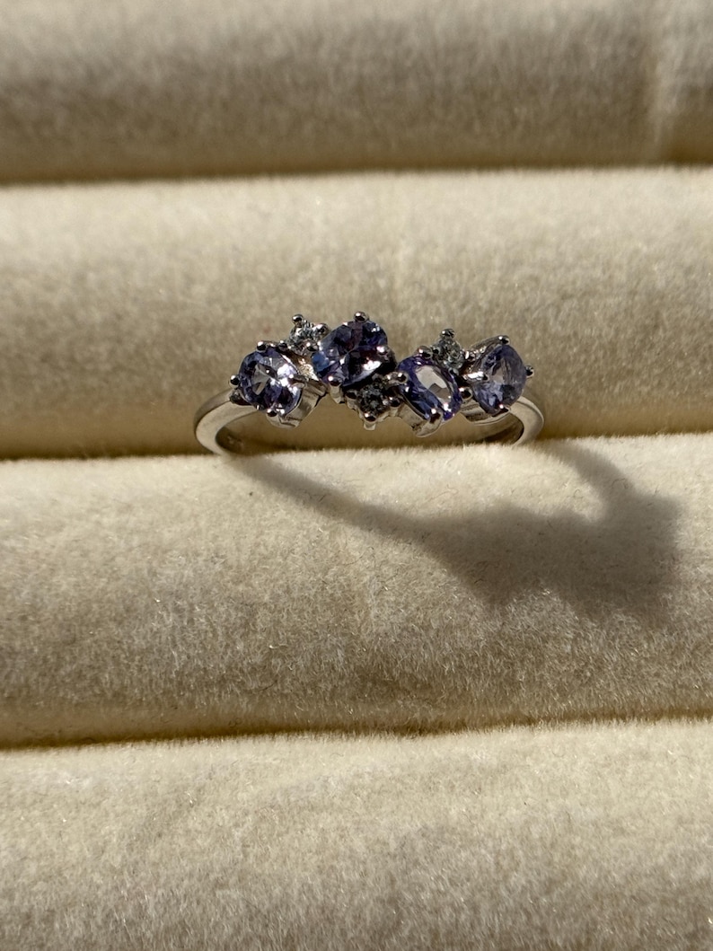 May include: A silver ring featuring five oval-shaped, light purple gemstones. The ring is set with smaller, clear accent stones. The ring is displayed on a light beige, textured surface.