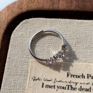May include: A silver ring with a row of small, light purple gemstones. The ring is set against a neutral-toned fabric inside a wooden tray. The ring's design is simple and elegant, with the gemstones adding a touch of color and sparkle.