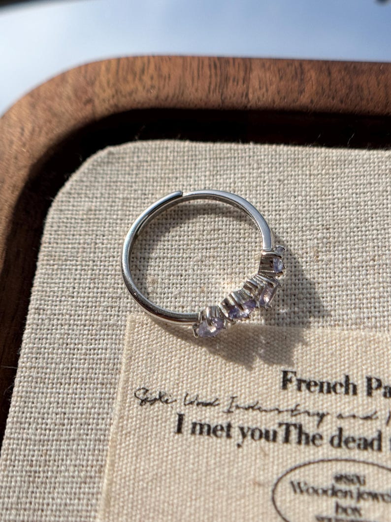 May include: A silver ring with a row of small, light purple gemstones. The ring is set against a neutral-toned fabric inside a wooden tray. The ring's design is simple and elegant, with the gemstones adding a touch of color and sparkle.