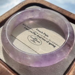 Op de afbeelding: Een doorschijnende lichtpaarse amethist armband rustend op een beige stoffen vierkant. De stof is voorzien van zwarte gedrukte tekst met French Paris, I met you The dead tree of spring en een ovaal logo voor Wooden jewelry.