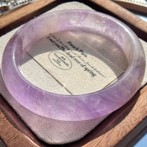 Peut inclure: Un bracelet jonc en améthyste violet clair translucide avec une finition lisse et polie. Le bracelet repose sur un carré de tissu blanc cassé à l'intérieur d'une boîte en bois. Le tissu porte le texte imprimé French Paris et I met you The dead tree of spring.