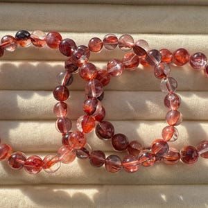 Op de afbeelding: Drie armbanden gemaakt van doorschijnende, roodbruine kralen. De ronde kralen zijn geregen op een transparant elastisch koord. De armbanden worden gepresenteerd op een lichtbeige oppervlak, waarbij het zonlicht de kralen benadrukt.