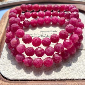 May include: Three ruby-colored beaded bracelets. Each bracelet is made of faceted, round beads. The bracelets are stacked on top of each other. The background includes the text "French Paris".