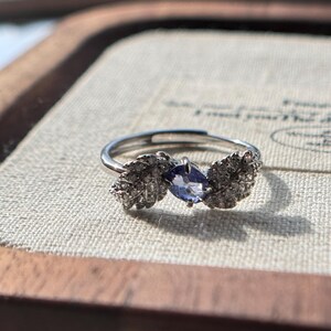 May include: A silver ring with a leaf design on each side of a blue gemstone. The ring is set on a light-colored surface, with a wooden frame in the background. The gemstone is oval-shaped and has a deep blue color.