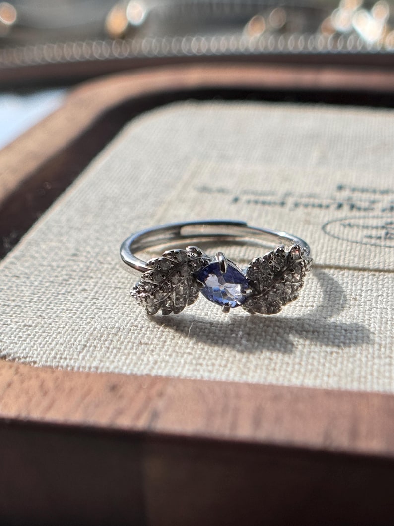 May include: A silver ring with a leaf design on each side of a blue gemstone. The ring is set on a light-colored surface, with a wooden frame in the background. The gemstone is oval-shaped and has a deep blue color.