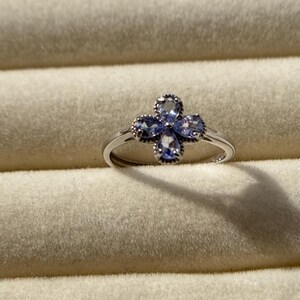 May include: A silver ring featuring a four-petal flower design. Each petal is adorned with a light blue gemstone. The ring is set against a neutral-toned background.
