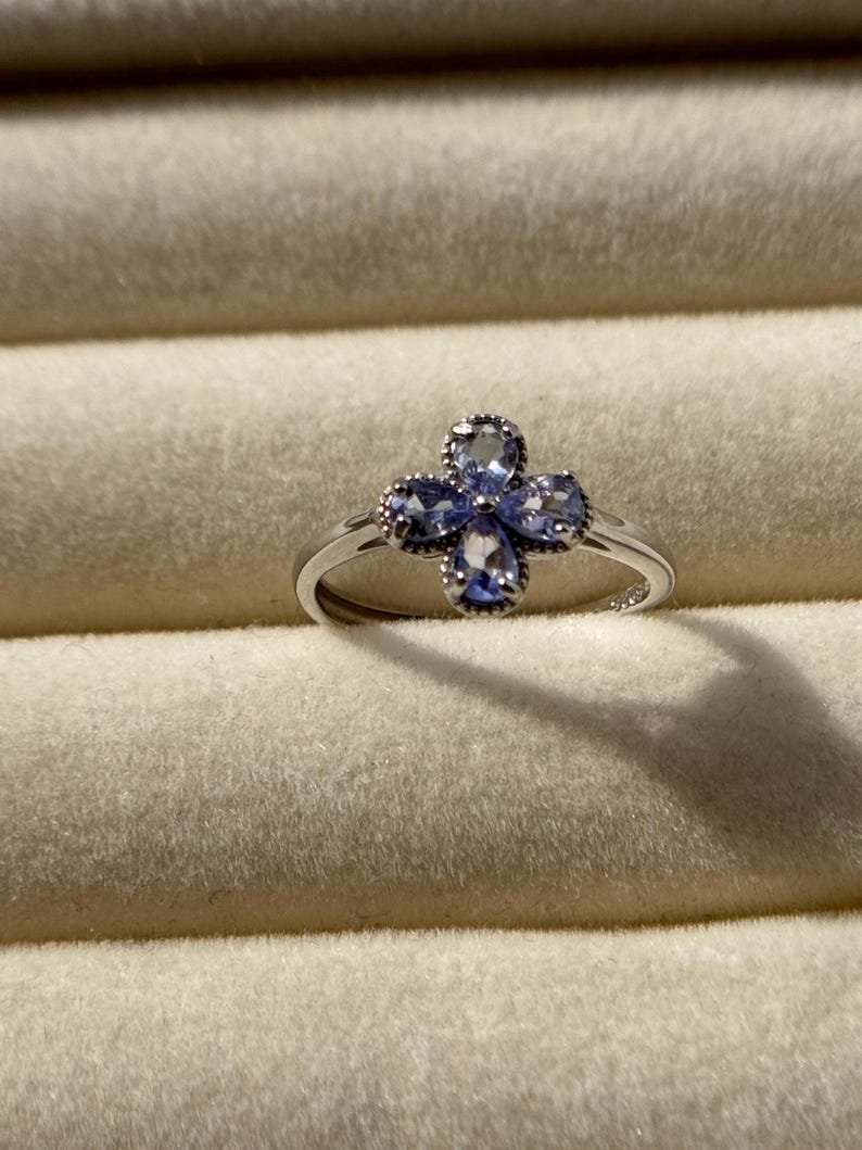 May include: A silver ring featuring a four-petal flower design. Each petal is adorned with a light blue gemstone. The ring is set against a neutral-toned background.