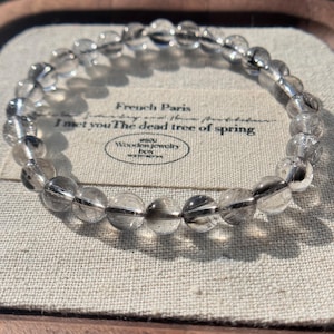 May include: A clear quartz bead bracelet with dark inclusions. The bracelet is made of round, polished beads strung together. The background includes text that reads "French Paris" and "I met you The dead tree of spring".