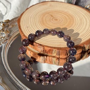 May include: A bracelet made of dark purple beads with translucent sections. The bracelet is presented on a wooden slice and a reflective surface, highlighting the natural colour variations and textures. The bracelet is a piece of jewellery.