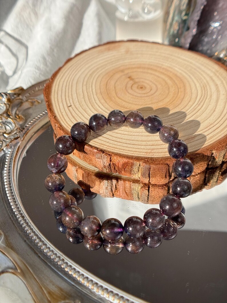 May include: A bracelet made of dark purple beads with translucent sections. The bracelet is presented on a wooden slice and a reflective surface, highlighting the natural colour variations and textures. The bracelet is a piece of jewellery.