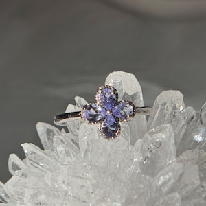 May include: A silver ring featuring a four-petal flower design. Each petal is composed of a light purple gemstone, with a delicate silver border. The ring is set against a backdrop of clear quartz crystals.