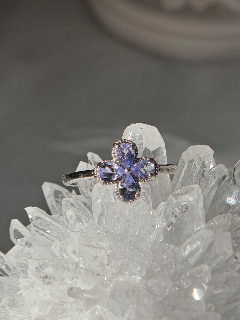May include: A silver ring featuring a four-petal flower design. Each petal is composed of a light purple gemstone, with a delicate silver border. The ring is set against a backdrop of clear quartz crystals.