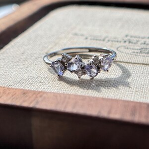 May include: A silver ring featuring a cluster of light purple gemstones and small clear stones. The ring's band is thin and polished, and the gemstones are arranged in a delicate, asymmetrical pattern. The ring is set against a neutral background.