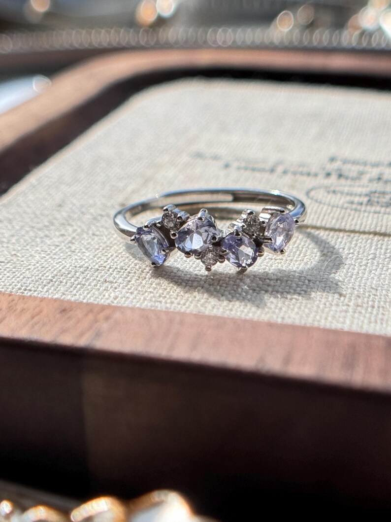 May include: A silver ring featuring a cluster of light purple gemstones and small clear stones. The ring's band is thin and polished, and the gemstones are arranged in a delicate, asymmetrical pattern. The ring is set against a neutral background.