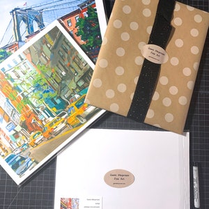Puede incluir: Un conjunto de tres impresiones enmarcadas de paisajes urbanos, que presentan un puente, edificios y coches. Las impresiones est&aacute;n empaquetadas en una bolsa de regalo de papel marr&oacute;n con lunares blancos y una cinta negra. La bolsa tiene una pegatina que dice "Gwen Meyerson Fine Art".
