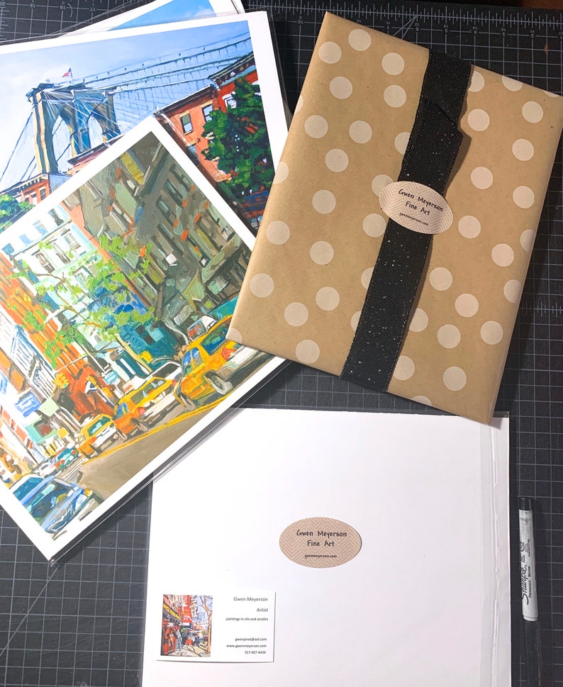 Puede incluir: Un conjunto de tres impresiones enmarcadas de paisajes urbanos, que presentan un puente, edificios y coches. Las impresiones est&aacute;n empaquetadas en una bolsa de regalo de papel marr&oacute;n con lunares blancos y una cinta negra. La bolsa tiene una pegatina que dice "Gwen Meyerson Fine Art".