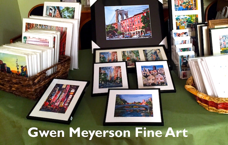 Puede incluir: Una mesa con una exposici&oacute;n de obras de arte enmarcadas que representan paisajes urbanos y puentes. Las obras est&aacute;n en marcos negros con paspart&uacute;s blancos. La mesa est&aacute; cubierta con un mantel verde. El texto "Gwen Meyerson Fine Art" es visible en la parte inferior de la imagen.
