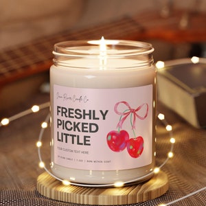 May include: A white candle in a clear glass jar with a pink label that says "Freshly Picked Little" and features a watercolor illustration of two red cherries with a pink ribbon. The candle is sitting on a wooden coaster.