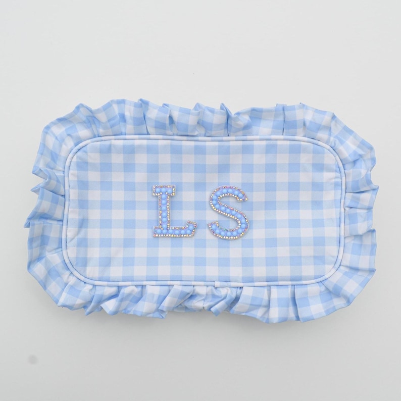 May include: A light blue and white gingham pattern pillow with a ruffled edge. The pillow features the embroidered initials "LS" in a decorative font, embellished with small, sparkling beads.