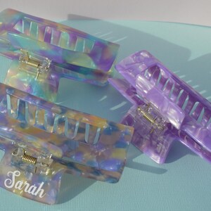 May include: Three iridescent hair claw clips in shades of purple, blue, and yellow. One clip has the name "Sarah" in a white script font. The clips are made of a marbled, translucent material.