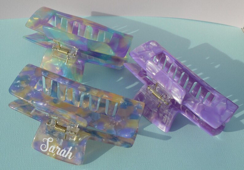 May include: Three iridescent hair claw clips in shades of purple, blue, and yellow. One clip has the name "Sarah" in a white script font. The clips are made of a marbled, translucent material.