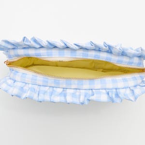 May include: A light blue and white gingham pattern cosmetic bag with a ruffled edge. The bag is open, revealing a yellow interior and a gold zipper. This accessory is perfect for storing makeup or toiletries.