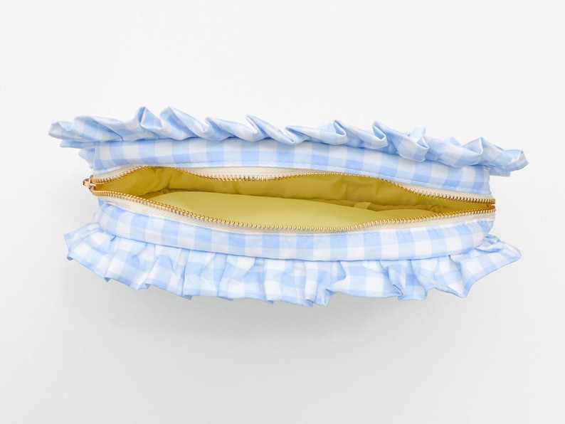 May include: A light blue and white gingham pattern cosmetic bag with a ruffled edge. The bag is open, revealing a yellow interior and a gold zipper. This accessory is perfect for storing makeup or toiletries.
