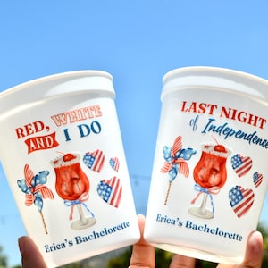 Könnte beinhalten: Zwei weiße Plastikbecher mit rot-weiß-blauen Designs. Auf einem Becher steht "RED, WHITE AND I DO" und auf dem anderen "LAST NIGHT of Independence". Jeder Becher zeigt eine Cocktail-Illustration, Windräder und Herzformen.