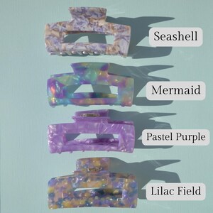May include: Four colorful hair claw clips in Seashell, Mermaid, Pastel Purple, and Lilac Field patterns. Each clip has a rectangular shape with a curved top and a metal clasp. The background is a light blue.