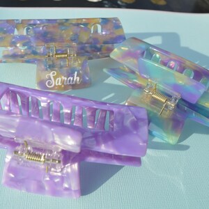 May include: Three colorful hair claw clips in shades of purple, blue, and yellow. One clip has the name "Sarah" in a cursive font. The clips have a marbled, iridescent appearance and a gold-colored spring mechanism.