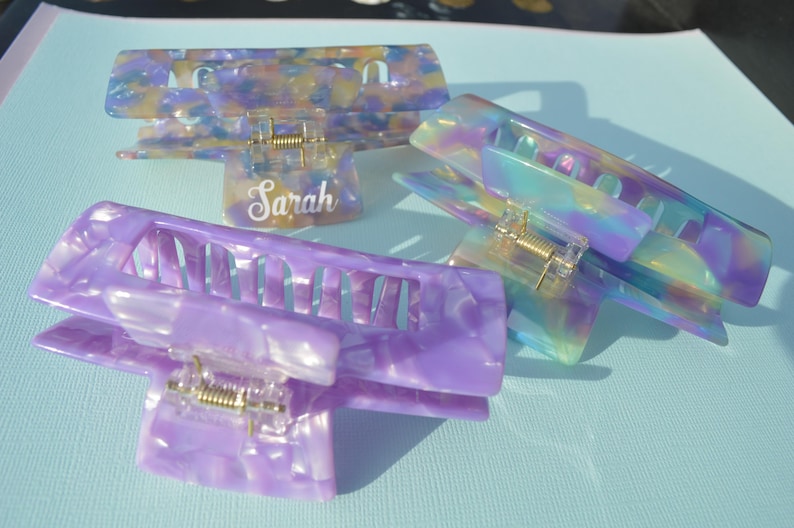 May include: Three colorful hair claw clips in shades of purple, blue, and yellow. One clip has the name "Sarah" in a cursive font. The clips have a marbled, iridescent appearance and a gold-colored spring mechanism.