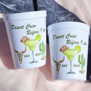 May include: Two white plastic cups with a desert-themed design. The cups feature a cactus, a skull, and a margarita glass. The text on the cups reads "Desert Crew Before I Do" and "EST 2025".