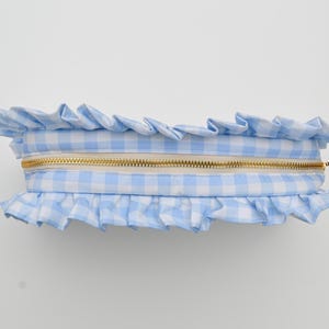 May include: A light blue and white gingham pattern cosmetic bag with a gold zipper. The bag is trimmed with ruffled fabric along the top and bottom edges. The bag is rectangular in shape.